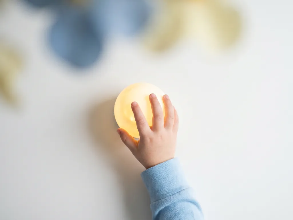 Klein kinderhandje boven een gloeiende bol op wit oppervlak, warme gele en lichtblauwe tinten, rustige minimalistische compositie.